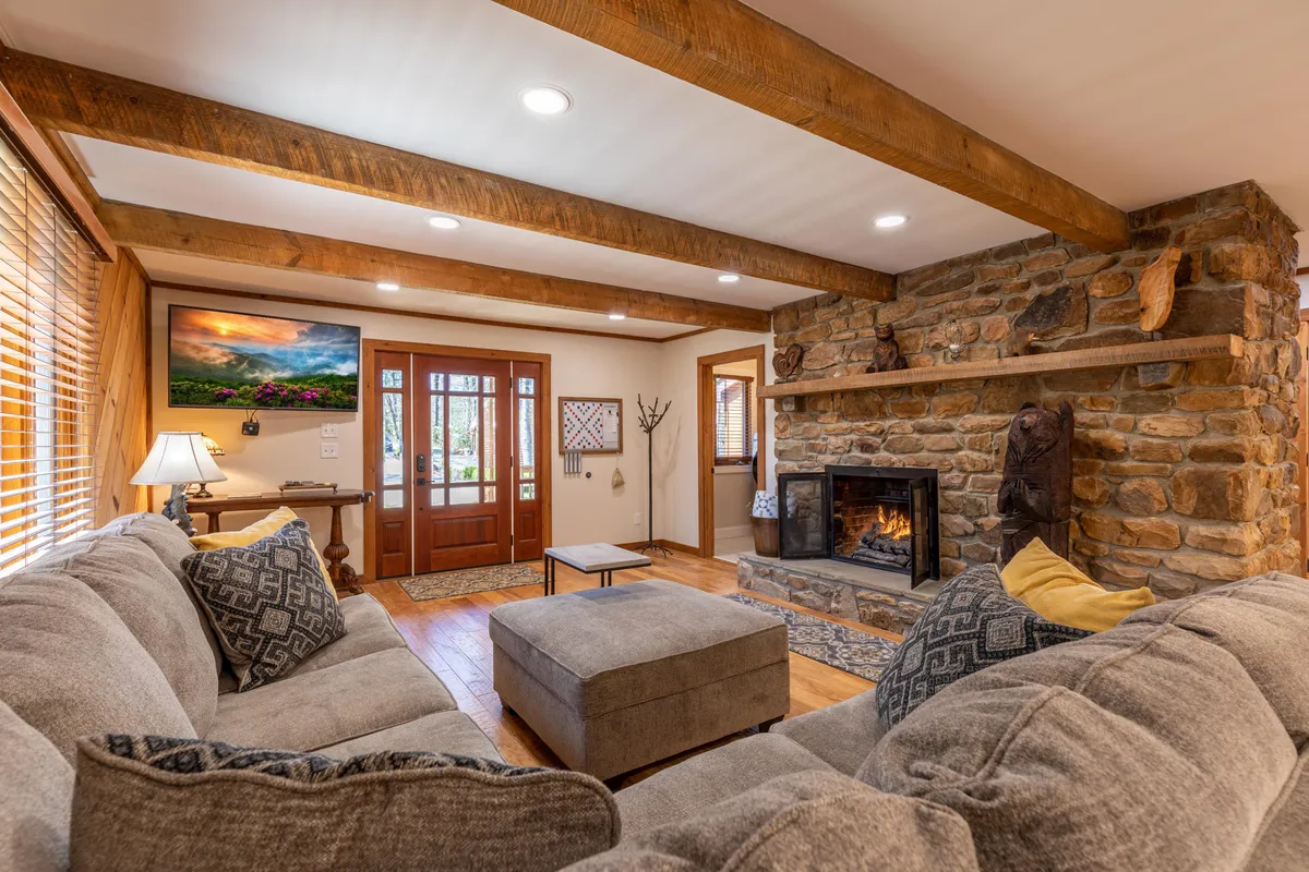 This photo features a bright interior space with a fireplace, large sectional sofa, rustic wooden beams, and the front door visible, creating a welcoming entrance atmosphere. The room is set up for relaxing and entertaining guests, reflecting a cozy and inviting mood.