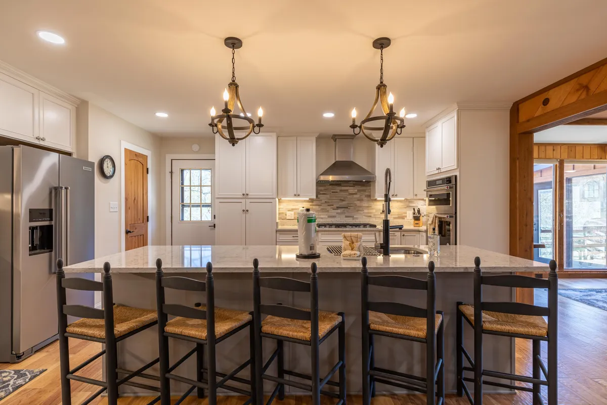 A spacious, modern kitchen with a large island, bar stool seating, elegant chandeliers, stainless steel appliances, and stylish backsplash tiles. The kitchen appears bright and inviting, perfect for both cooking and gathering.