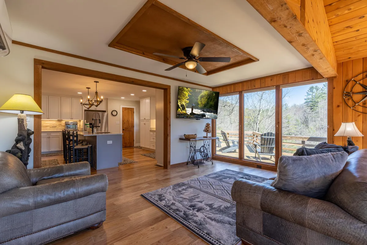 Here, the main living room is shown with comfortable sofas, large windows letting in natural light and views of nature, a modern ceiling fan, and tasteful decorative touches. The space feels airy, cozy, and ideal for unwinding or socializing.