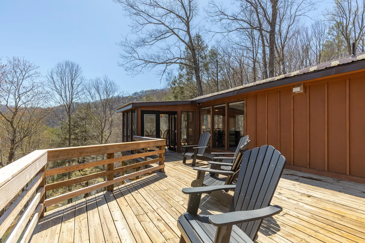 A spacious wooden deck with Adirondack chairs positioned to face a breathtaking mountain view. The deck is surrounded by greenery, and the morning sunlight softly illuminates the area, creating a tranquil atmosphere ideal for relaxation.