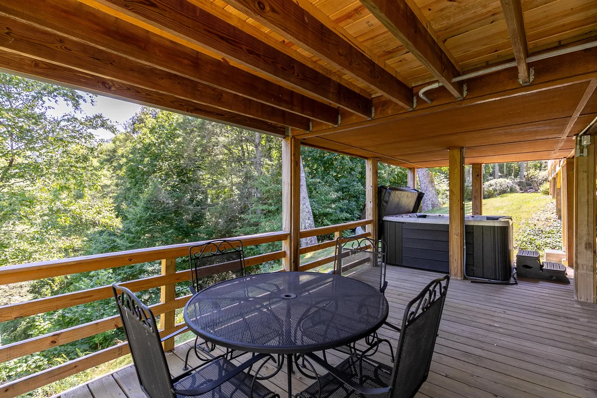 An outdoor dining area on a wooden deck, surrounded by lush greenery, with a hot tub nearby. The setup offers an inviting space for al fresco meals, relaxation, and soaking in natural views, blending indoor comfort with outdoor leisure.
