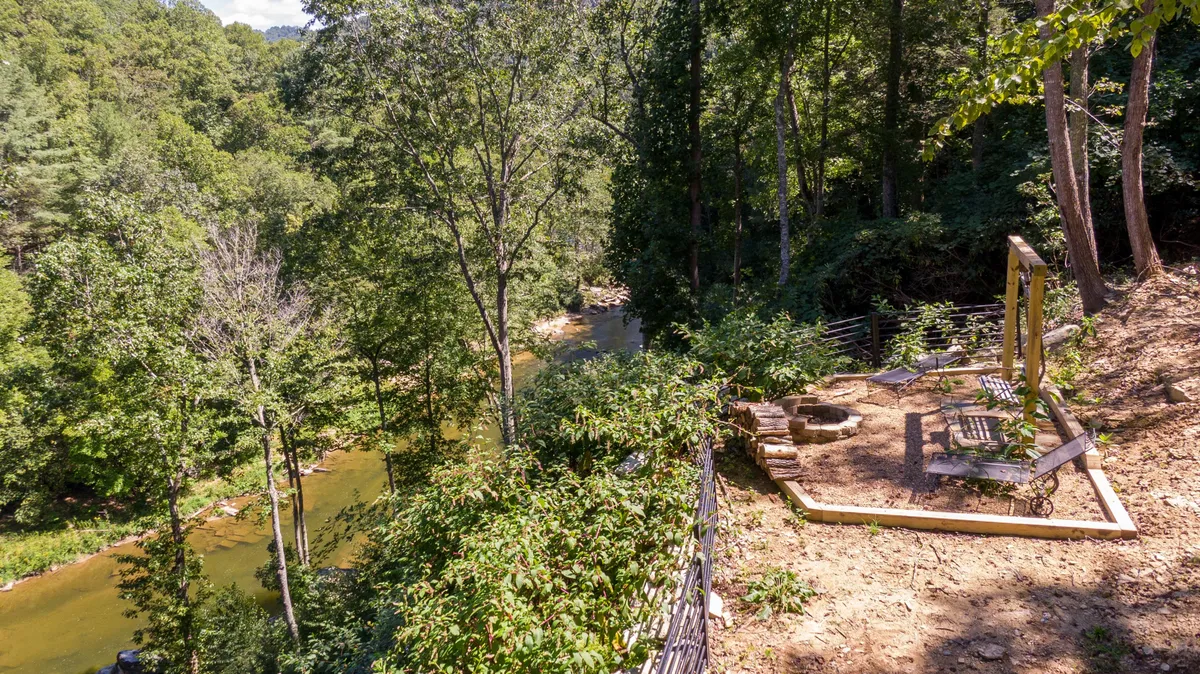 A scenic riverside seating area features Adirondack chairs arranged around a fire pit, surrounded by trees and overlooking the flowing river. The image exudes a sense of relaxation and togetherness, with the fire pit inviting families to gather for s'mores after a day of adventure. The natural backdrop emphasizes the expansive, private grounds and the seamless connection between the property and outdoor fun.