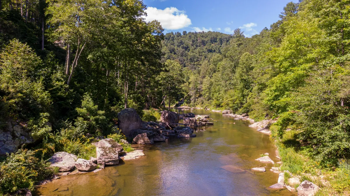 This image captures a calm section of the river as it winds through lush greenery, with rock formations and mature trees lining the banks. The scene is serene and inviting, evoking the sense of private riverfront access. The tranquil water suggests opportunities for fishing, skipping stones, or launching tubes, while the natural setting highlights the adventure and exploration potential right in the backyard.