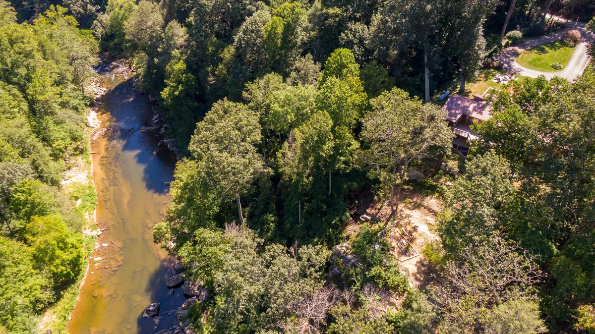 A serene view of the river bordered by dense trees with the house visible in the background. The tranquil water, lush greenery, and hints of wildlife create a peaceful environment ideal for fishing, hiking, picnicking, and wildlife observation.