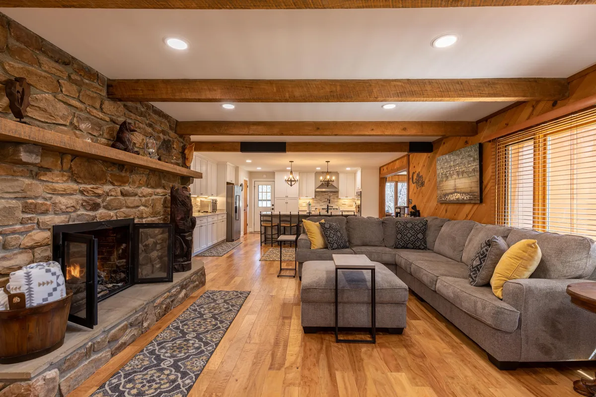 This image showcases a spacious living room with a prominent stone fireplace, a large sectional sofa for group seating, exposed wooden beams, a decorative rug, and a view into the open kitchen area in the background. The mood is warm and welcoming, perfect for gatherings and relaxation.