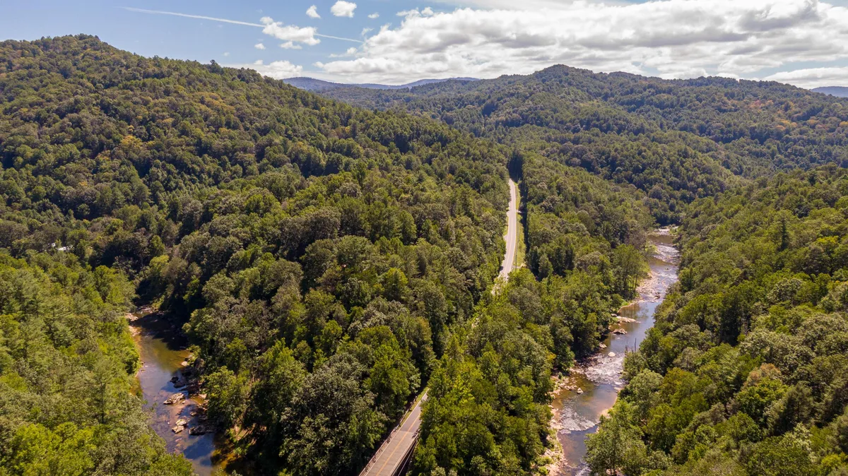A sweeping view of a mountainous landscape with a winding road, river, and dense forest. The scene offers a sense of expansive wilderness, perfect for hiking, nature photography, bird watching, and kayaking. The river and forested areas evoke the tranquility and adventure of the natural world right outside the property.