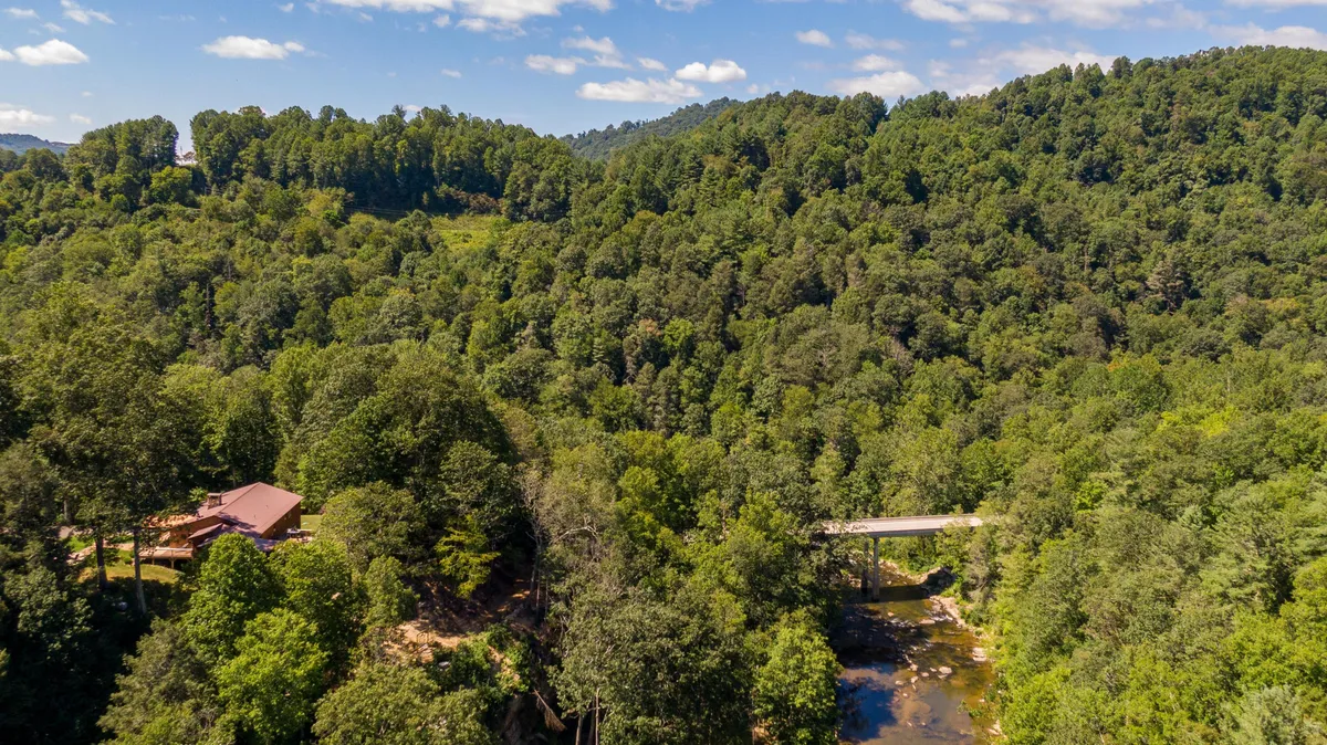 A tranquil exterior view shows a forested landscape with a gently flowing stream, a small rustic bridge, and a cozy cabin nestled among the trees. The scene feels peaceful and highlights the natural beauty that surrounds the home.