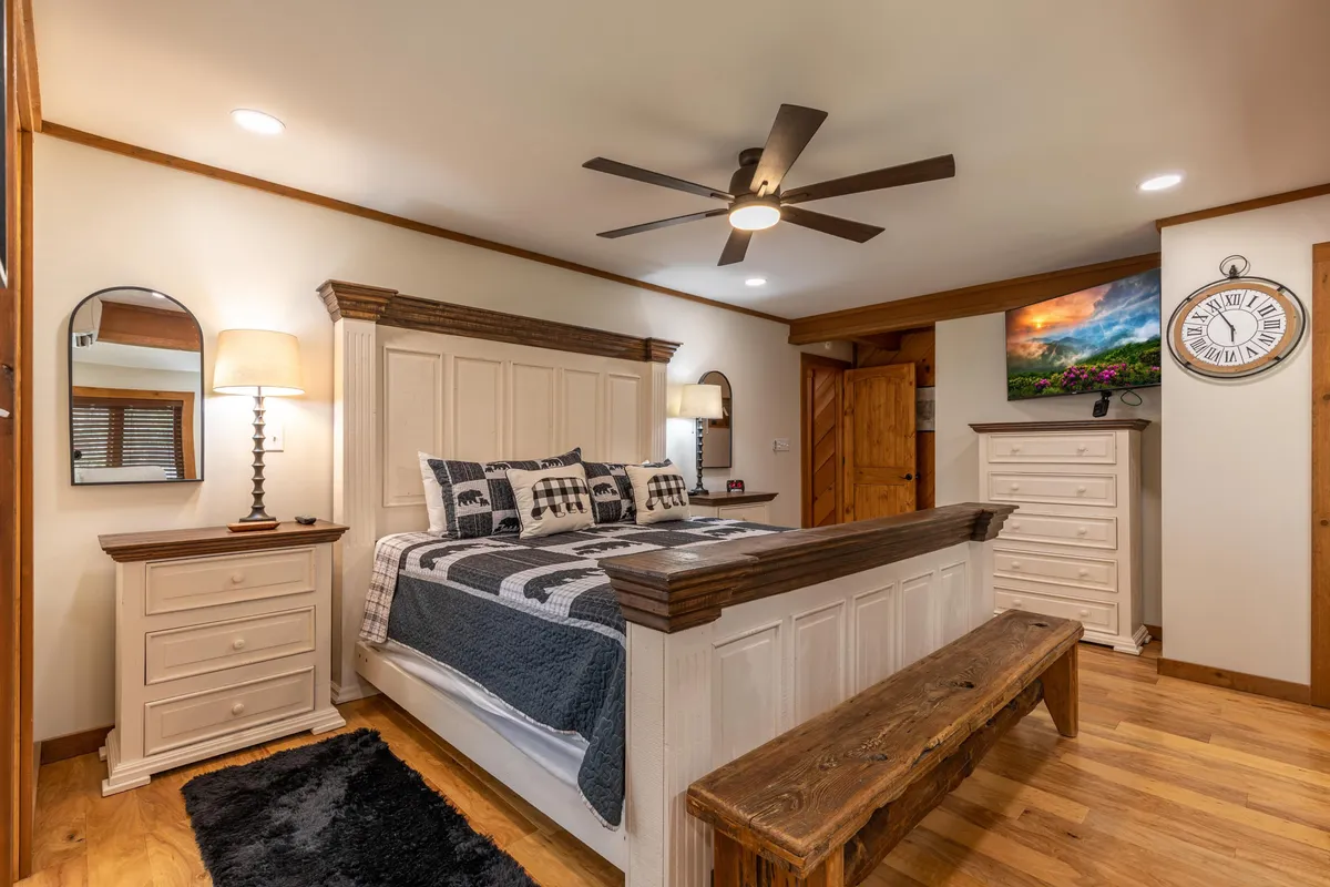 A king bedroom featuring stylish bedding, a wooden bench at the foot of the bed, landscape wall art, modern ceiling fan, and nightstands with lamps. The room has a serene and inviting atmosphere, ideal for relaxation and restful nights.