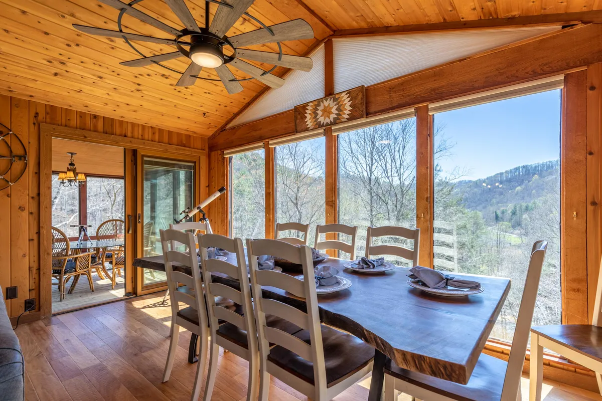 A cozy dining area featuring a large wooden table, ample seating for eight, expansive windows providing natural light, and a wood-paneled ceiling with a ceiling fan. The ambiance is perfect for family meals or entertaining guests.