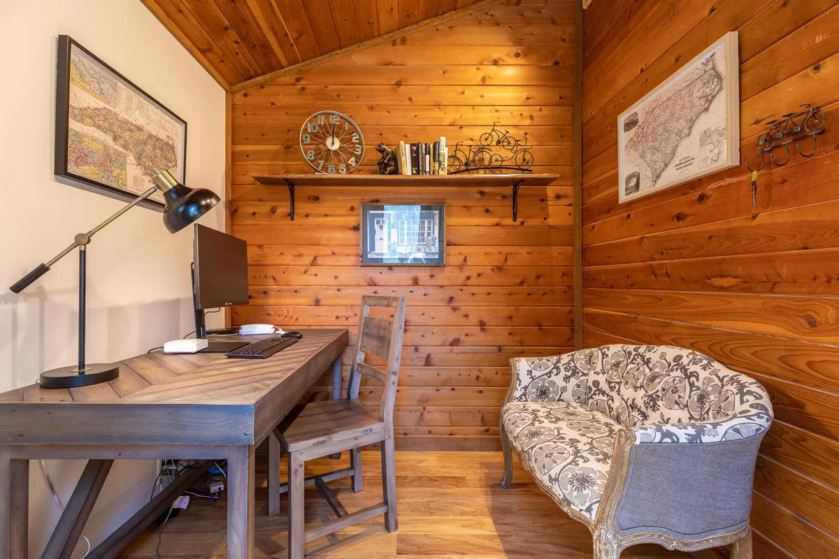 A modern and comfortable office space featuring a wooden desk, decorative chair, a shelf with books, and a wall clock. The workspace is well-lit and inviting, ideal for productive work sessions or quiet reading.