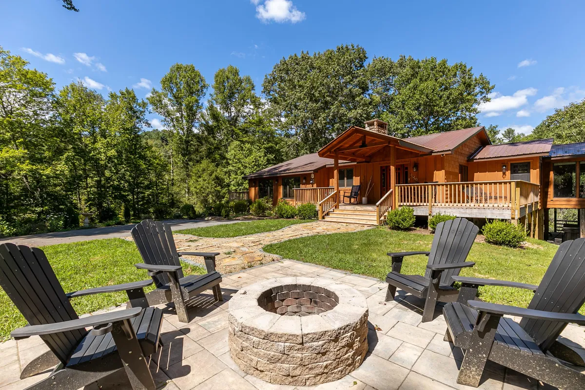 The hero image shows a charming wooden house nestled among tall trees, with a cozy fire pit surrounded by Adirondack chairs in the foreground. The scene captures the inviting outdoor gathering space, perfect for relaxing evenings, and hints at sunset views and the tranquility of the natural setting. The trees and lush greenery frame the house, conveying a sense of privacy and immersion in nature.