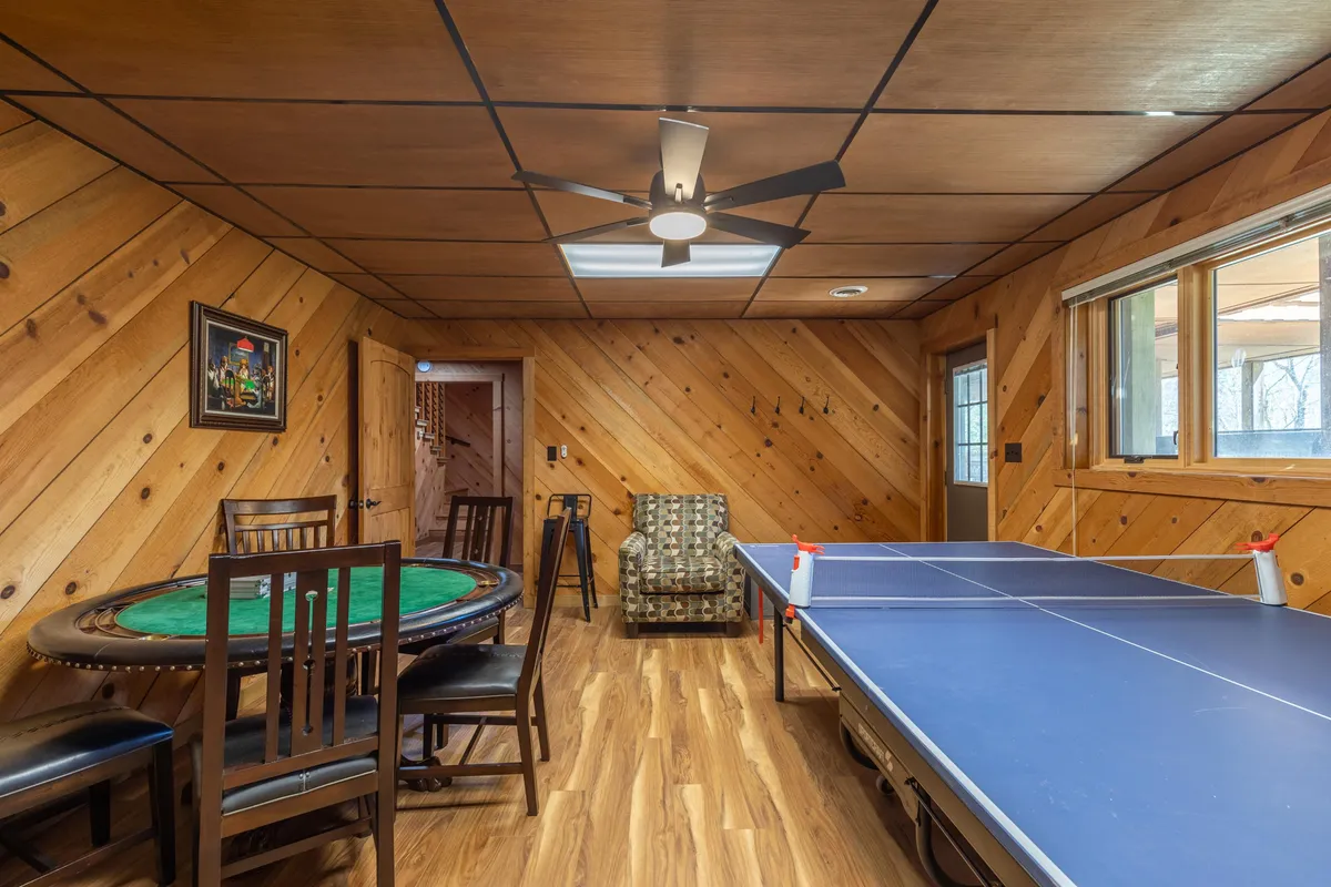 A game room featuring a ping pong table and poker table, with large windows letting in natural light and wooden walls enhancing the recreational feel. The setup invites friendly competition and social breaks.