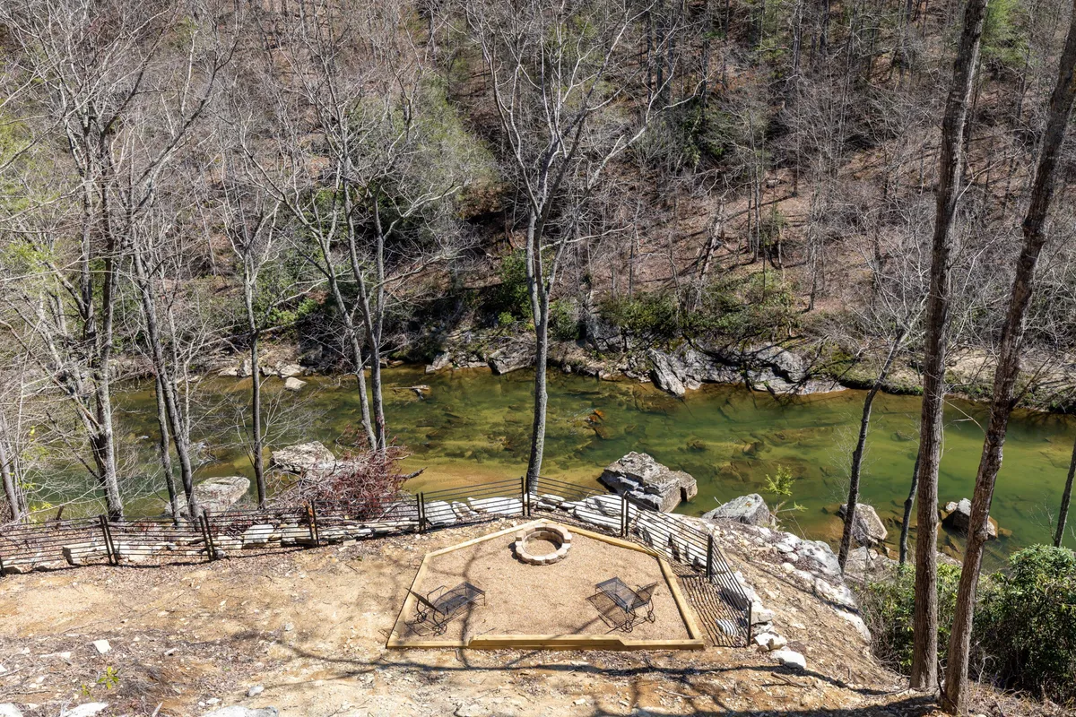 A tranquil riverside fire pit area with comfortable seating, river views, and tree-lined surroundings. The scene invites guests to gather for marshmallow roasts, storytelling, and stargazing by the water.