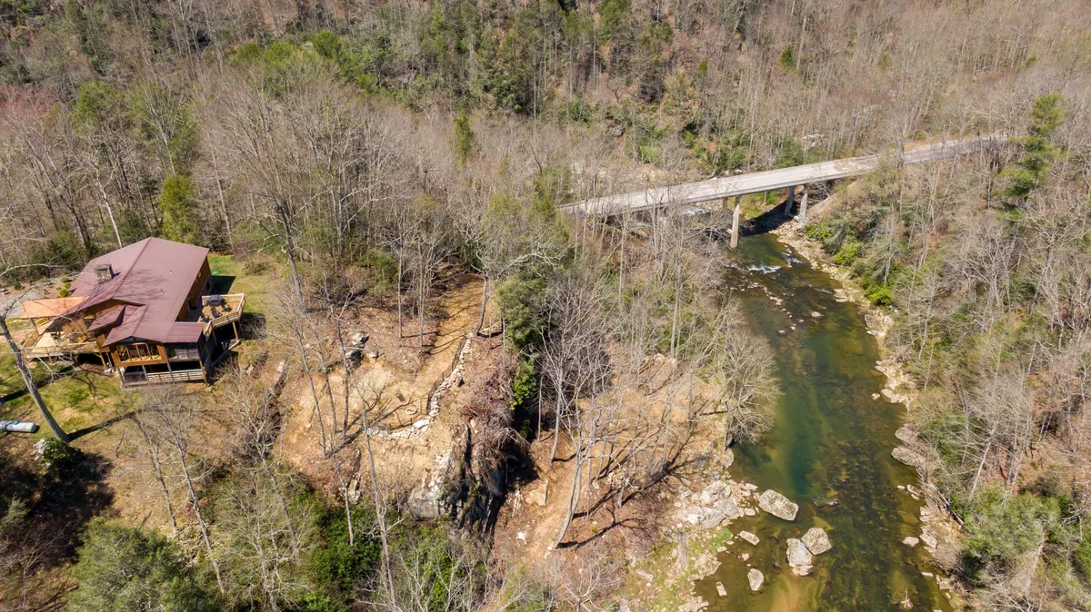 A view of a rustic cabin nestled by the flowing river, surrounded by a dense forest and with a bridge in the background. The scene highlights both the natural beauty of the property and its built features, showing that the home is truly immersed in the outdoors. The image suggests a private trail, opportunities for wildlife spotting, and the adventure of exploring expansive, wooded acreage.